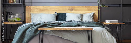 Panorama of wooden bench in front of bed with green blanket in grey bedroom interior. Real photo