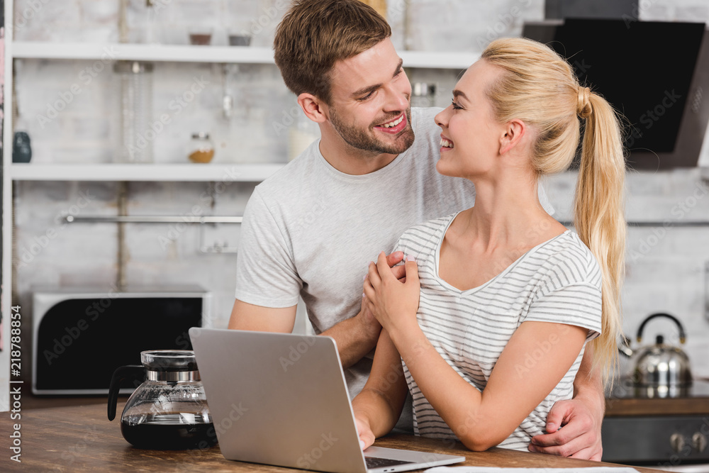 happy boyfriend hugging girlfriend in kitchen while she working with laptop and they looking at each other happy boyfriend hugging girlfriend in kitchen while she working with laptop and they looking at each other