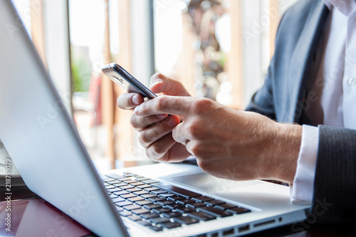 Businessman in black suit using mobile smart phone and working on laptop computer, browsing internet and writing on paper notebook in modern office. Man working on electronics devices with copy space