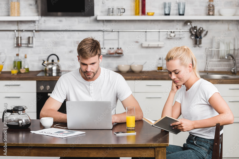 young man using laptop and smiling woman reading book while sitting together at kitchen table young man using laptop and smiling woman reading book while sitting together at kitchen table