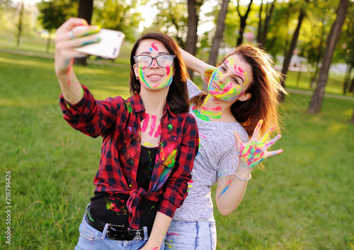 two young pretty girls smeared with colored fingers paint themselves on the camera smartphone  grimace and make selfi
