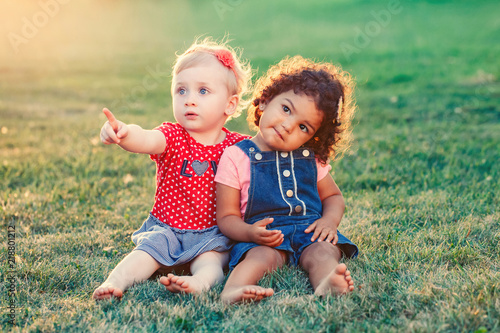 Group portrait of two cute adorable girls toddlers children sitting together. White Caucasian and latin hispanic babies hugging outside in park. Friendship and best friends forever concept.