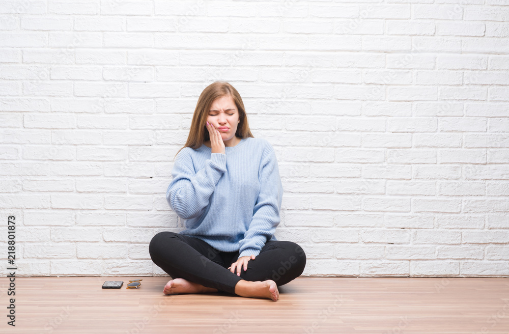 Young adult woman sitting on the floor in autumn over white brick wall touching mouth with hand with painful expression because of toothache or dental illness on teeth. Dentist concept. Young adult woman sitting on the floor in autumn over white brick wall touching mouth with hand with painful expression because of toothache or dental illness on teeth. Dentist concept.