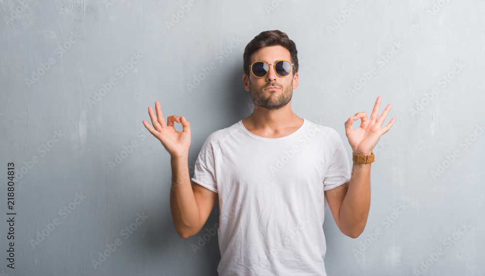 Handsome young man over grey grunge wall wearing sunglasses relax and smiling with eyes closed doing meditation gesture with fingers. Yoga concept. Handsome young man over grey grunge wall wearing sunglasses relax and smiling with eyes closed doing meditation gesture with fingers. Yoga concept.