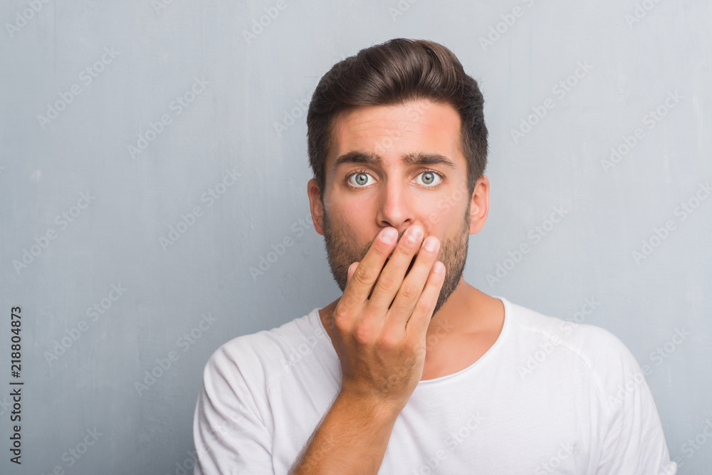 Handsome young man over grey grunge wall cover mouth with hand shocked with shame for mistake, expression of fear, scared in silence, secret concept Handsome young man over grey grunge wall cover mouth with hand shocked with shame for mistake, expression of fear, scared in silence, secret concept