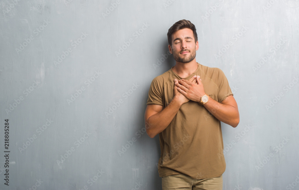 Handsome young man over grey grunge wall smiling with hands on chest with closed eyes and grateful gesture on face. Health concept.