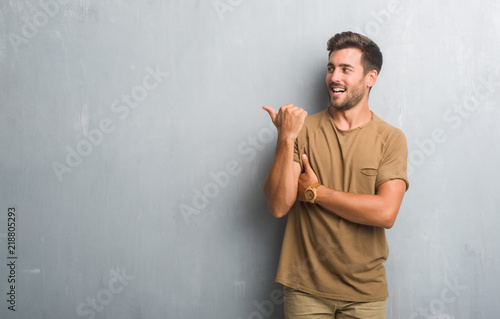 Handsome young man over grey grunge wall smiling with happy face looking and pointing to the side with thumb up.