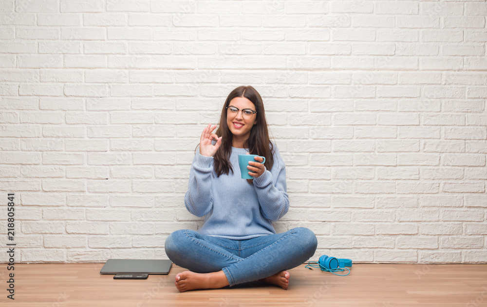 Young brunette woman sitting on the floor over white brick wall drinking coffee doing ok sign with fingers, excellent symbol Young brunette woman sitting on the floor over white brick wall drinking coffee doing ok sign with fingers, excellent symbol