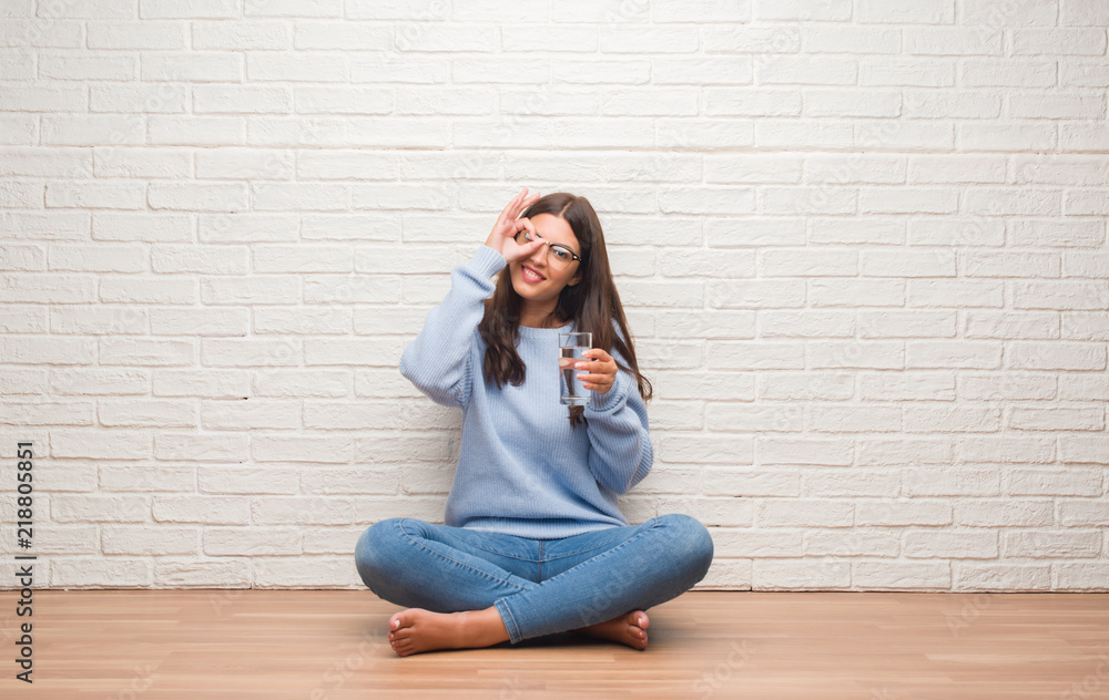 Young brunette woman sitting on the floor drinking glass of water with happy face smiling doing ok sign with hand on eye looking through fingers Young brunette woman sitting on the floor drinking glass of water with happy face smiling doing ok sign with hand on eye looking through fingers