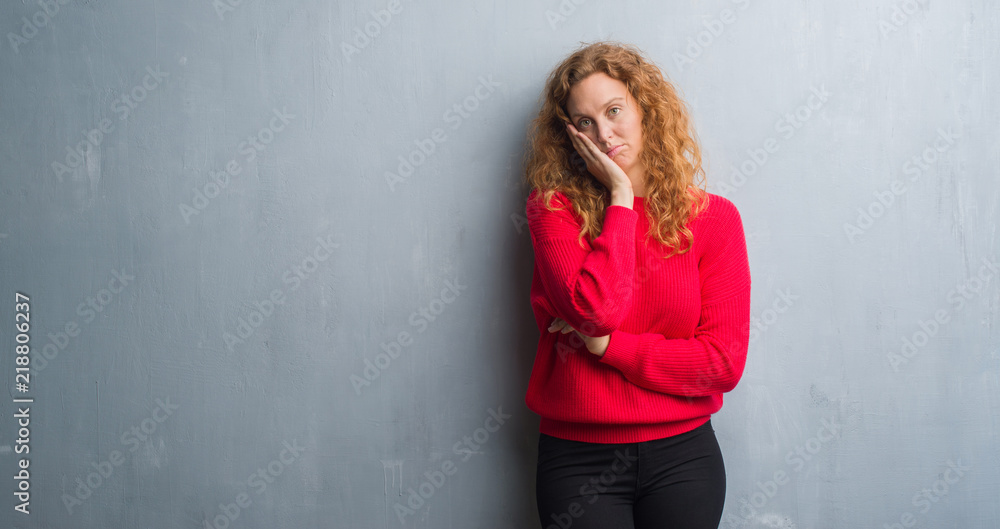 Young redhead woman over grey grunge wall wearing red sweater thinking looking tired and bored with depression problems with crossed arms.