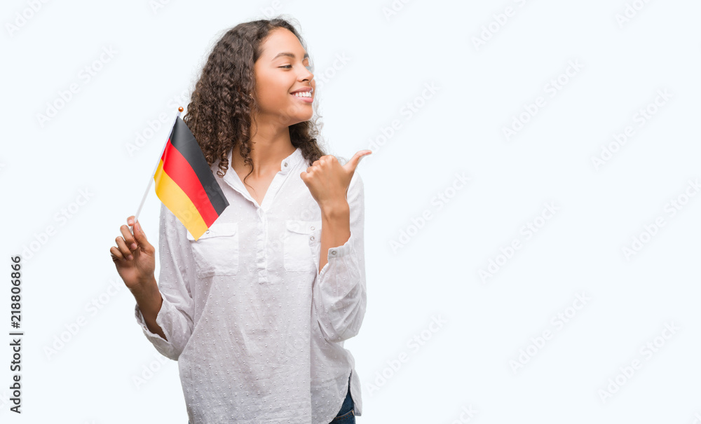 Young hispanic woman holding flag of Germany pointing and showing with thumb up to the side with happy face smiling Young hispanic woman holding flag of Germany pointing and showing with thumb up to the side with happy face smiling