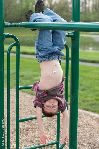 Fotografija Young boy with gastric g-tube hanging upside down from monkey bars on jungle gym playground at park