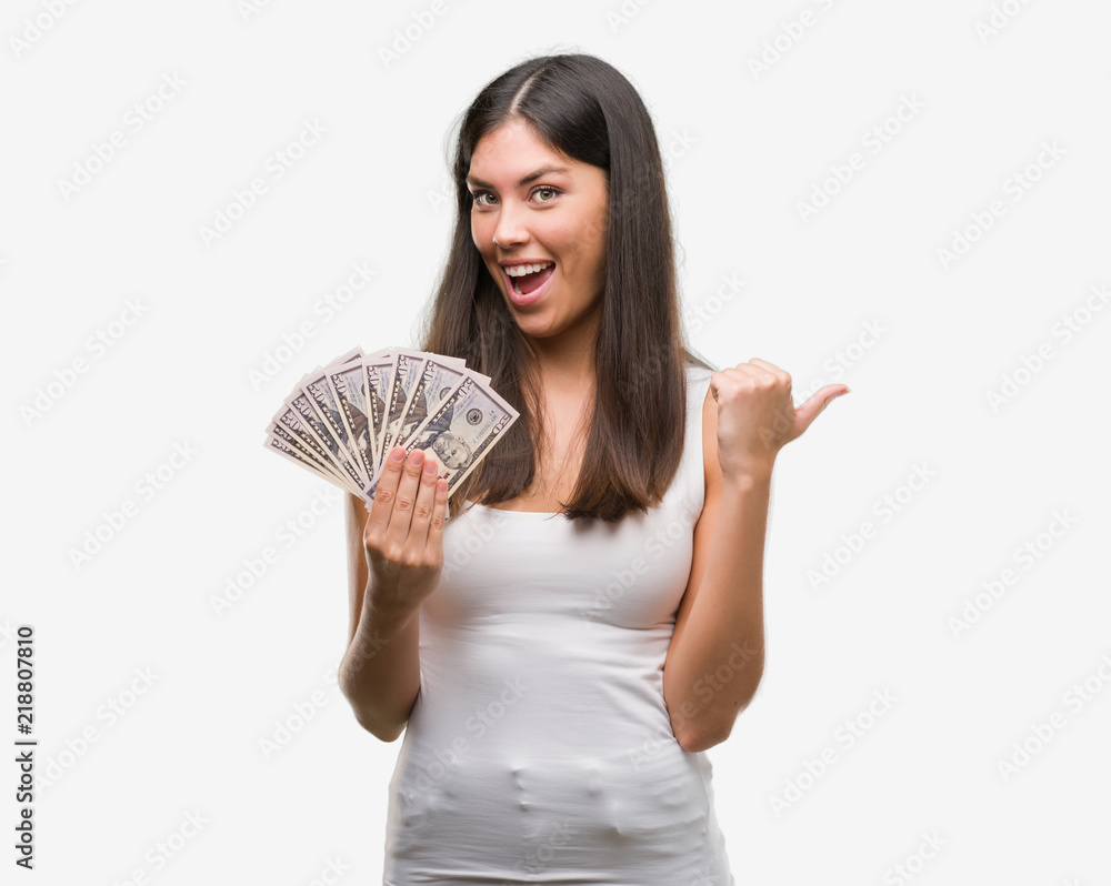 Young hispanic woman holding dollars pointing and showing with thumb up to the side with happy face smiling Young hispanic woman holding dollars pointing and showing with thumb up to the side with happy face smiling