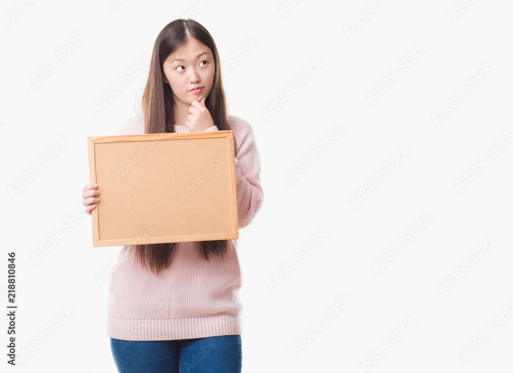 Young Chinese woman over isolated background holding corkboard serious face thinking about question, very confused idea Young Chinese woman over isolated background holding corkboard serious face thinking about question, very confused idea
