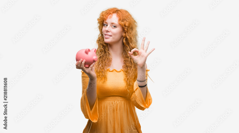 Young redhead woman holding piggy bank doing ok sign with fingers, excellent symbol