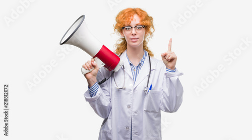 Young redhead doctor woman holding megaphone surprised with an idea or question pointing finger with happy face, number one
