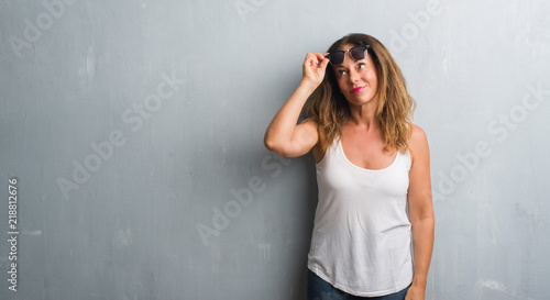 Middle age hispanic woman over grey wall wearing sunglasses smiling looking side and staring away thinking. © Krakenimages.com