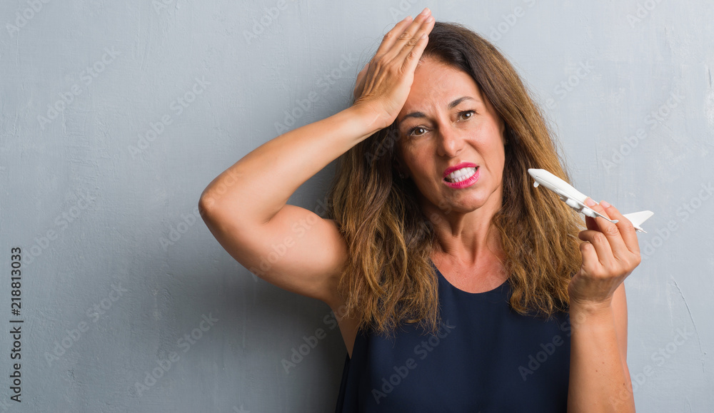 Middle age hispanic woman standing over grey grunge wall holding airplane stressed with hand on head, shocked with shame and surprise face, angry and frustrated. Fear and upset for mistake.