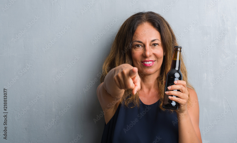 Middle age hispanic woman standing over grey grunge wall holding beer bottle pointing with finger to the camera and to you, hand sign, positive and confident gesture from the front Middle age hispanic woman standing over grey grunge wall holding beer bottle pointing with finger to the camera and to you, hand sign, positive and confident gesture from the front