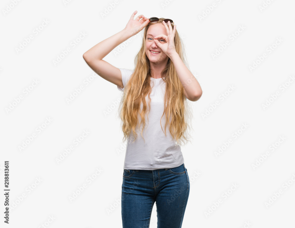 Blonde teenager woman wearing sunglasses with happy face smiling doing ok sign with hand on eye looking through fingers Blonde teenager woman wearing sunglasses with happy face smiling doing ok sign with hand on eye looking through fingers