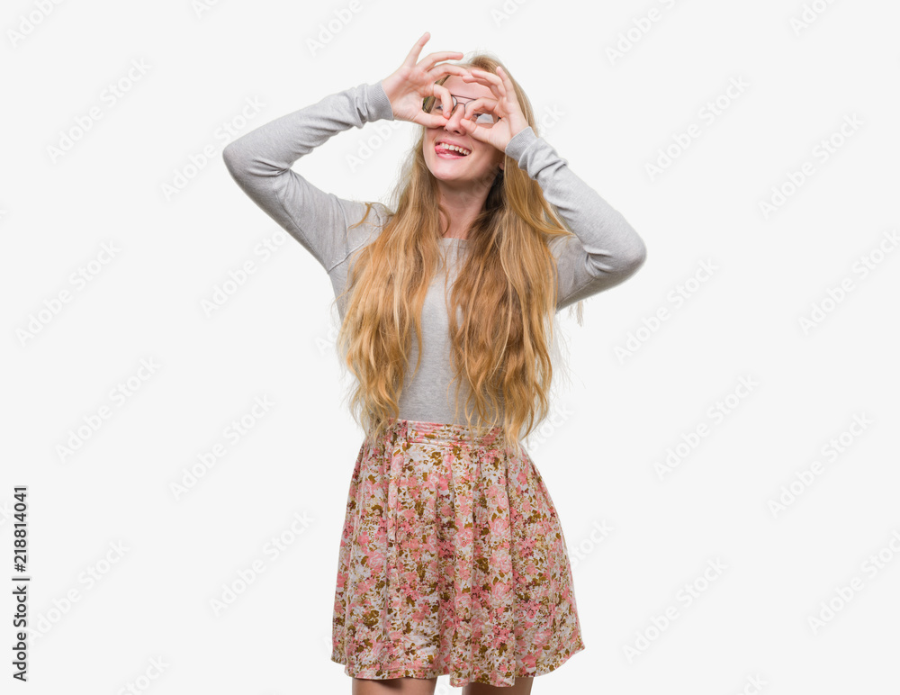 Blonde teenager woman wearing flowers skirt doing ok gesture like binoculars sticking tongue out, eyes looking through fingers. Crazy expression. Blonde teenager woman wearing flowers skirt doing ok gesture like binoculars sticking tongue out, eyes looking through fingers. Crazy expression.