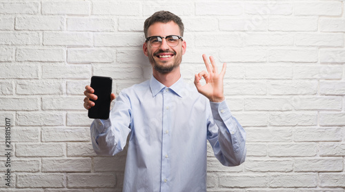 Young adult man talking on the phone standing over white brick wall doing ok sign with fingers, excellent symbol