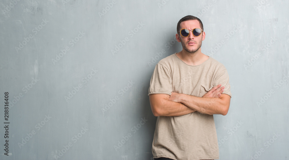 Young caucasian man over grey grunge wall wearing sunglasses skeptic and nervous, disapproving expression on face with crossed arms. Negative person. Young caucasian man over grey grunge wall wearing sunglasses skeptic and nervous, disapproving expression on face with crossed arms. Negative person.