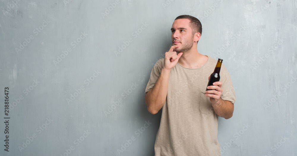 Young caucasian man over grey grunge wall holding bottle beer serious face thinking about question, very confused idea