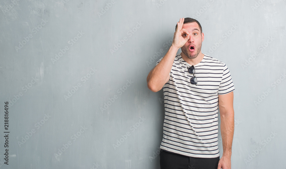 Young caucasian man over grey grunge wall doing ok gesture shocked with surprised face, eye looking through fingers. Unbelieving expression. Young caucasian man over grey grunge wall doing ok gesture shocked with surprised face, eye looking through fingers. Unbelieving expression.
