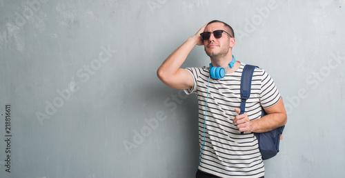 Young caucasian student man over grey grunge wall wearing headphones and backpack stressed with hand on head, shocked with shame and surprise face, angry and frustrated. Fear and upset for mistake.