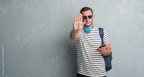 Young caucasian student man over grey grunge wall wearing headphones using smartphone with open hand doing stop sign with serious and confident expression, defense gesture