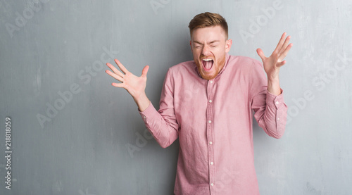 Young redhead man over grey grunge wall wearing pink shirt celebrating mad and crazy for success with arms raised and closed eyes screaming excited. Winner concept