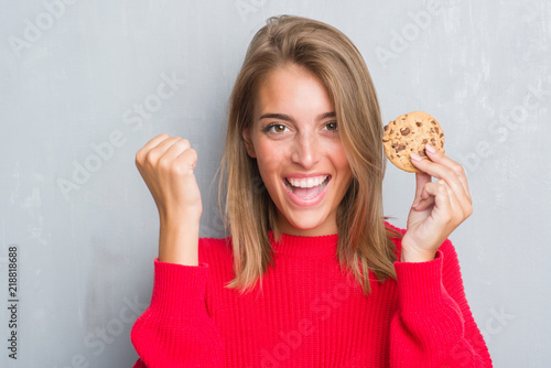 Beautiful young woman over grunge grey wall eating chocolate chip cooky screaming proud and celebrating victory and success very excited  cheering emotion