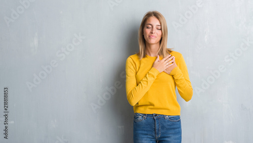 Beautiful young woman standing over grunge grey wall smiling with hands on chest with closed eyes and grateful gesture on face. Health concept.