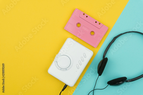 top view of a pink audio cassette tape with a white portable cassette player and a black headphone on the yellow and blue background