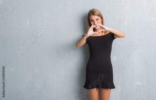 Beautiful young woman standing over grunge grey wall wearing elegant dress smiling in love showing heart symbol and shape with hands. Romantic concept. © Krakenimages.com