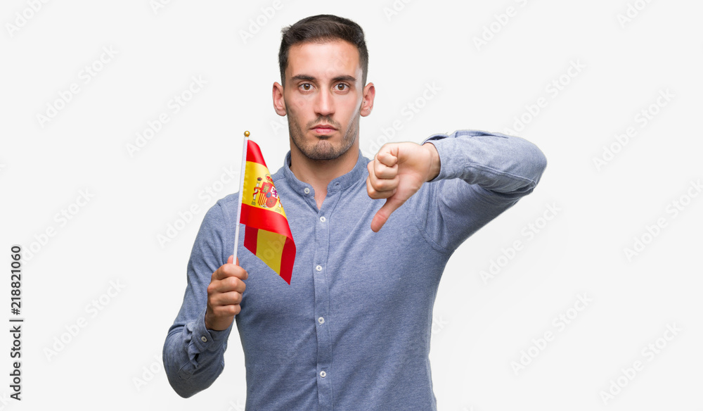 Handsome young man holding a flag of Spain with angry face, negative sign showing dislike with thumbs down, rejection concept Handsome young man holding a flag of Spain with angry face, negative sign showing dislike with thumbs down, rejection concept