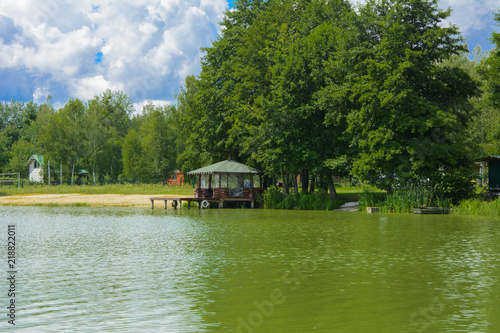 A beautiful image of a landscape from the center of a river surrounded by trees and reeds on the shore against a blue sky in the clouds. Wooden gazebo on beach. Reflection, water, tourist destination