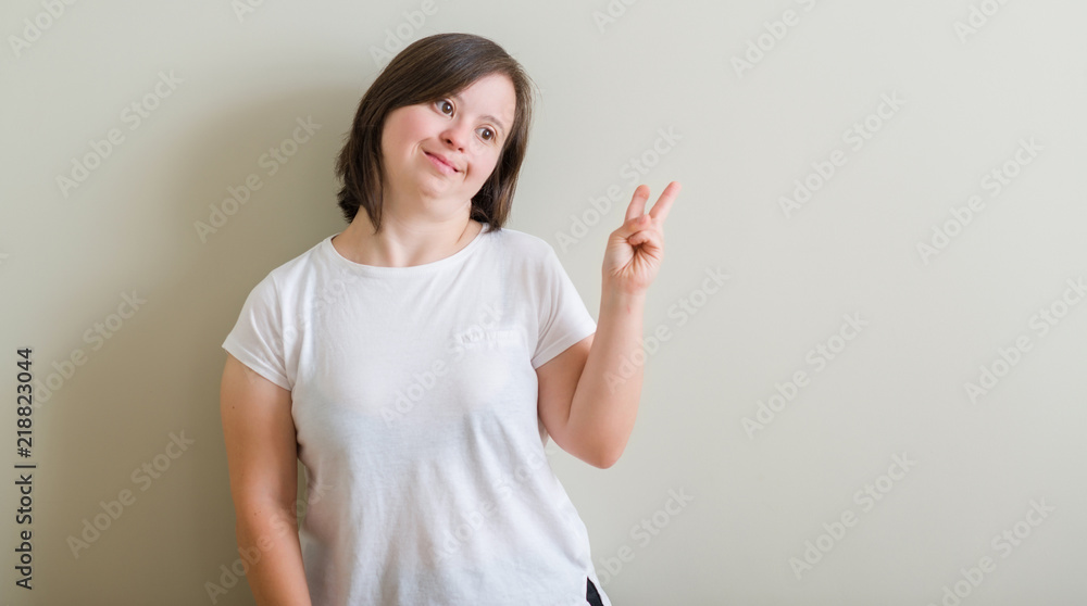 Down syndrome woman standing over wall smiling with happy face winking at the camera doing victory sign. Number two. Down syndrome woman standing over wall smiling with happy face winking at the camera doing victory sign. Number two.