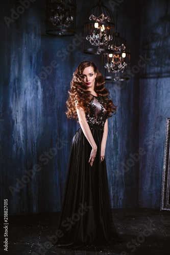 Young woman with long curly hair and makeup in evening long luxury dress, posing in a dark interior room. fashion beauty portrait