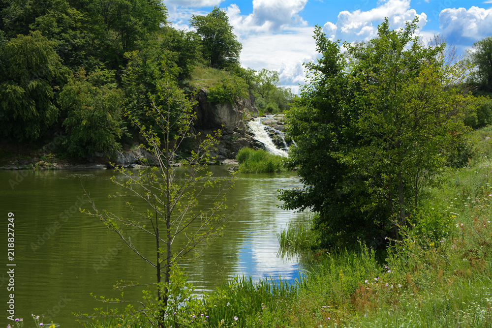 The waterfall on the river flows through and over the rocks covered with lichen and moss against a background of green vegetation and a blue sky. The waterfall on the river flows through and over the rocks covered with lichen and moss against a background of green vegetation and a blue sky.