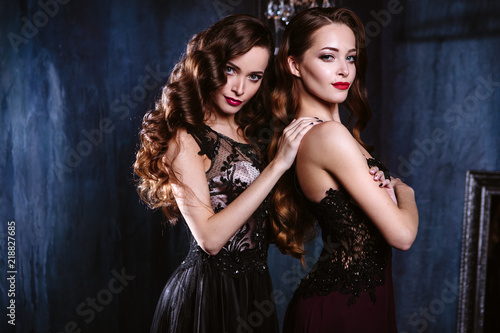 Twins young women in evening dresses, fashion beauty portrait in dark interior