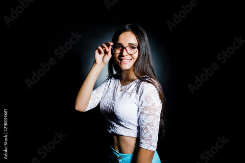 Portrait of an attractive young woman in white top and blue pants posing with her glasses in the dark.