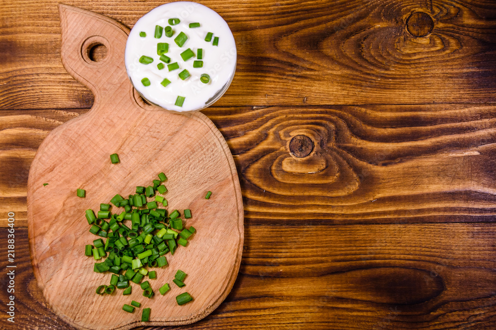 Glass bowl with sour cream and cutting board with chopped green onion on wooden table. Top view Glass bowl with sour cream and cutting board with chopped green onion on wooden table. Top view