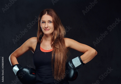 Smiling sportive woman dressed in sportswear wearing boxing gloves posing in a studio. Isolated on dark textured background.