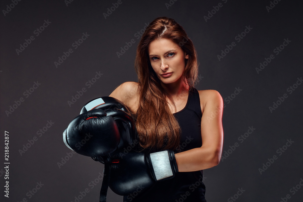 Sportive woman dressed in sportswear wearing boxing gloves holds protective helmet. Isolated on dark background.