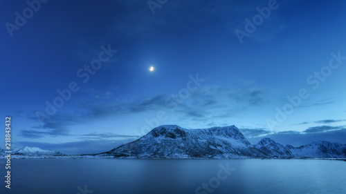 Snow covered mountains against blue sky with clouds and moon in winter at night in Lofoten islands, Norway. Arctic landscape with sea, snowy rocks, moonlight, reflection in water. Beautiful fjord photo