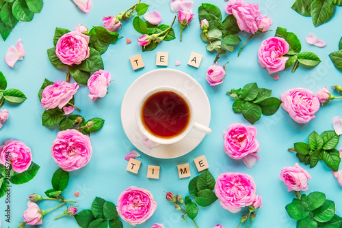 Top view creative layout with Tea time lettering with wooden blocks, cup of hot tea and fresh pink tea rose flowers, buds, petals, leaves on the blue background. Flat lay. Selective focus. Copy space.