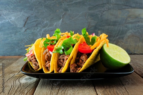 Hard shelled tacos with ground beef, lettuce, tomatoes and cheese. Group on plate with a dark background.