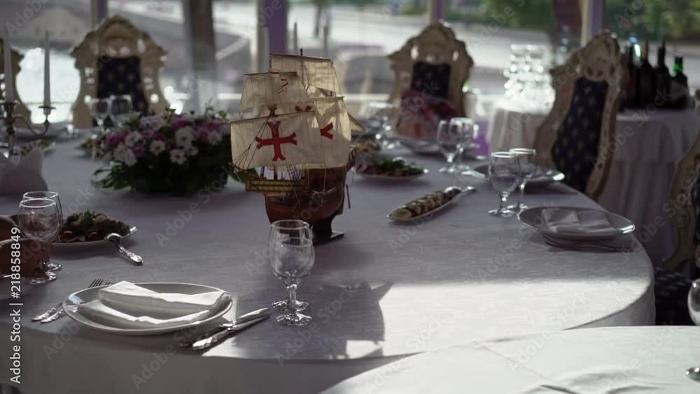 Plate, cloth napkin, knife and fork on white cloth on table in luxury restaurant decorated with model of old wooden ship, candles and flowers composition Plate, cloth napkin, knife and fork on white cloth on table in luxury restaurant decorated with model of old wooden ship, candles and flowers composition